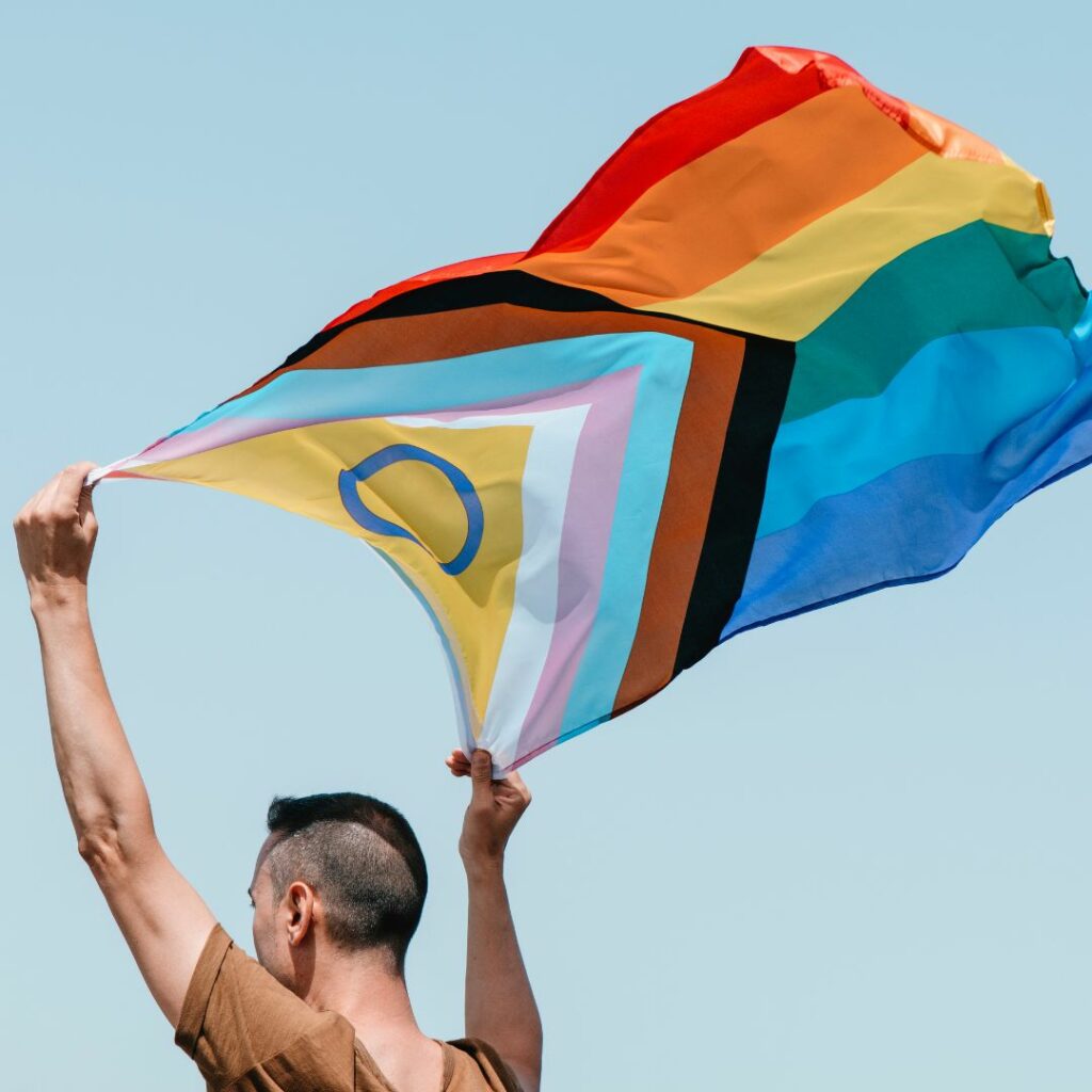 Person med irokesfrisyr håller en progressiv Pride-flagga över huvudet, flaggan vajar i vinden.