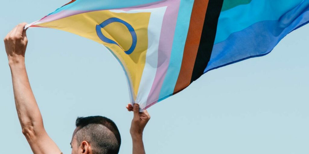 A person with mohawk hair is flying a progressive Pride flag over their head, the flag swaying in the wind.