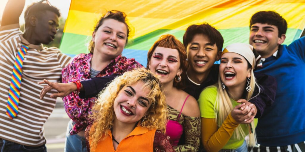 A diverse group of young people are smiling at the camera, in front of a Pride flag.