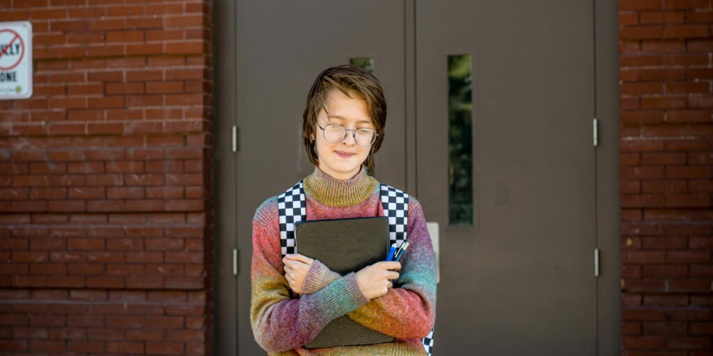 A young person with a rainbow-coloured knitted polo stands in front of the doors to a brick building, calmly looking downwards. They hold a folder and two ballpens in their hands.